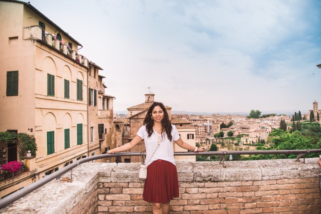 13 Best Towns In Tuscany 26 A woman in a white dress with long dark hair standing at a scenic viewpoint overlooking the medieval terracotta rooftops and towers of Siena spreading across the Tuscan hills, with green cypress trees and rolling countryside visible in the background