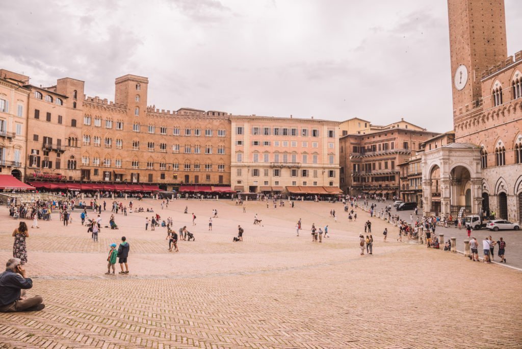 15 Amazing Day Trips From Florence, Italy 14 Wide view of Piazza del Duomo in Siena showing the exterior of Siena Cathedral with its distinctive black and white striped marble facade, Gothic arches and detailed stone sculptures, with tourists gathered in the square and a cloudy grey sky above