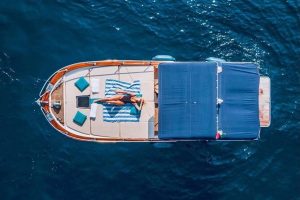 5 Best Private Boat Tours Of Capri 12 Aerial view of a person lounging on a private boat deck with blue and white striped cushions, surrounded by the tranquil blue waters of the ocean, illustrating a luxurious and relaxing boating experience.