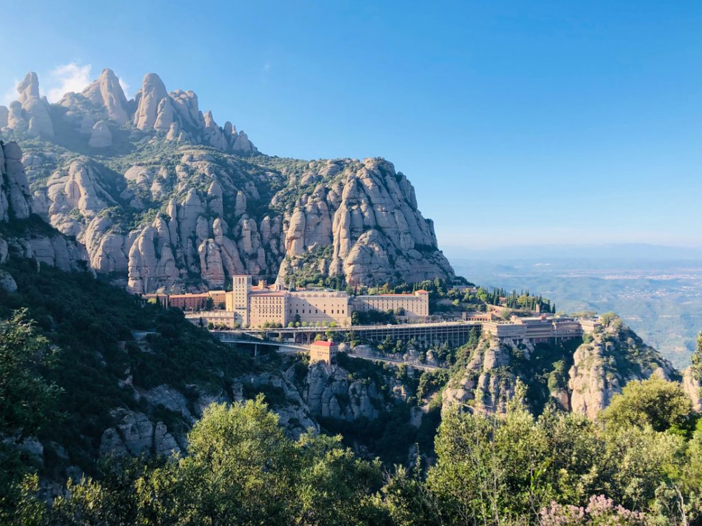 34 Incredible Weekend Trips From Barcelona 12 The Montserrat Monastery nestled in the multi-peaked mountain range of Catalonia, Spain, with a panoramic view of rugged cliffs, verdant foliage, and a clear blue sky.
