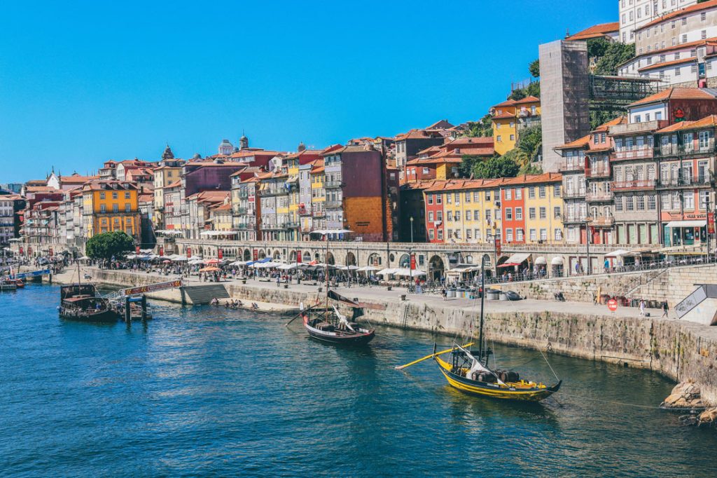 34 Incredible Weekend Trips From Barcelona 41 The vibrant Ribeira District of Porto, Portugal, with traditional Rabelo boats on the Douro River and colorful historic buildings lining the waterfront promenade under a clear blue sky.