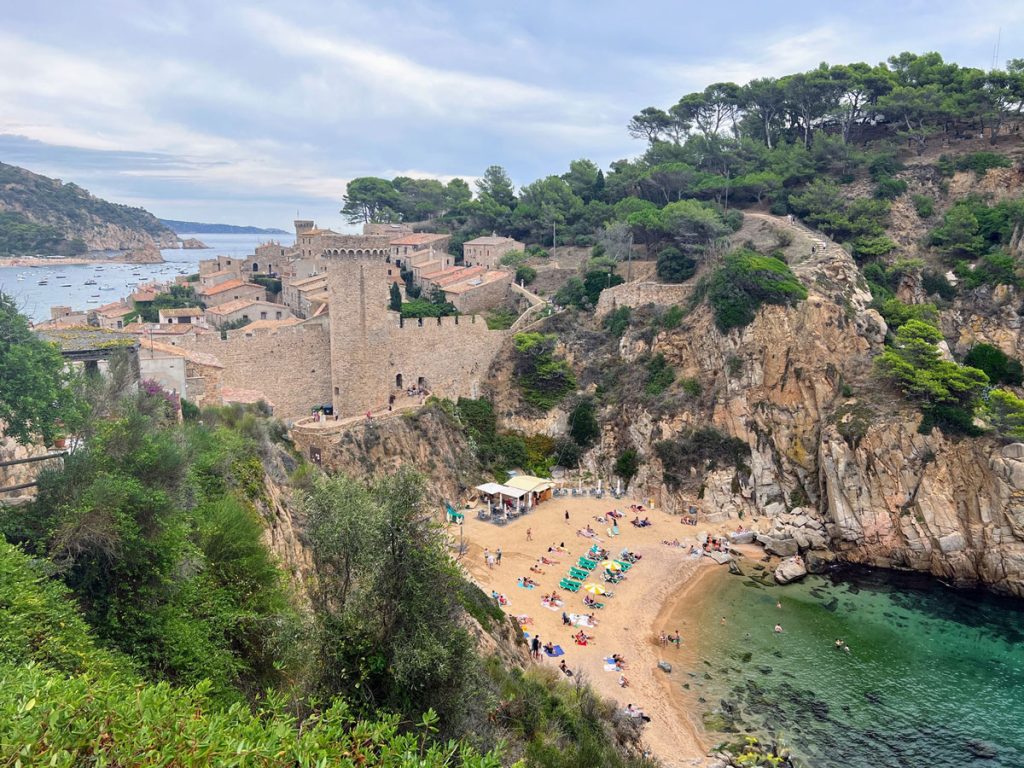 34 Incredible Weekend Trips From Barcelona 18 A scenic view of Tossa de Mar beach in Spain, showcasing the historic town walls and medieval towers adjacent to a cliff-lined cove with beachgoers on the sand and clear turquoise waters.