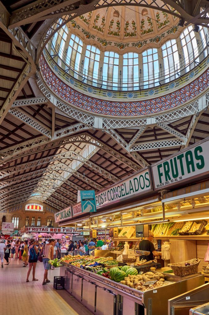 34 Incredible Weekend Trips From Barcelona 25 Inside view of Valencia's bustling local market, showcasing vendors at colorful fruit and vegetable stalls with the text 'Frutas' prominently displayed, under an ornate, dome-shaped ceiling with intricate metalwork.