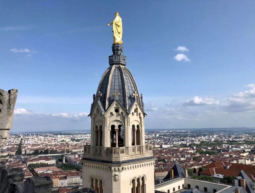34 Incredible Weekend Trips From Barcelona 38 The golden statue of Mary towers atop the roof of the Basilica of Notre-Dame de Fourvière, with a panoramic view of Lyon's cityscape stretching into the horizon under a clear sky.