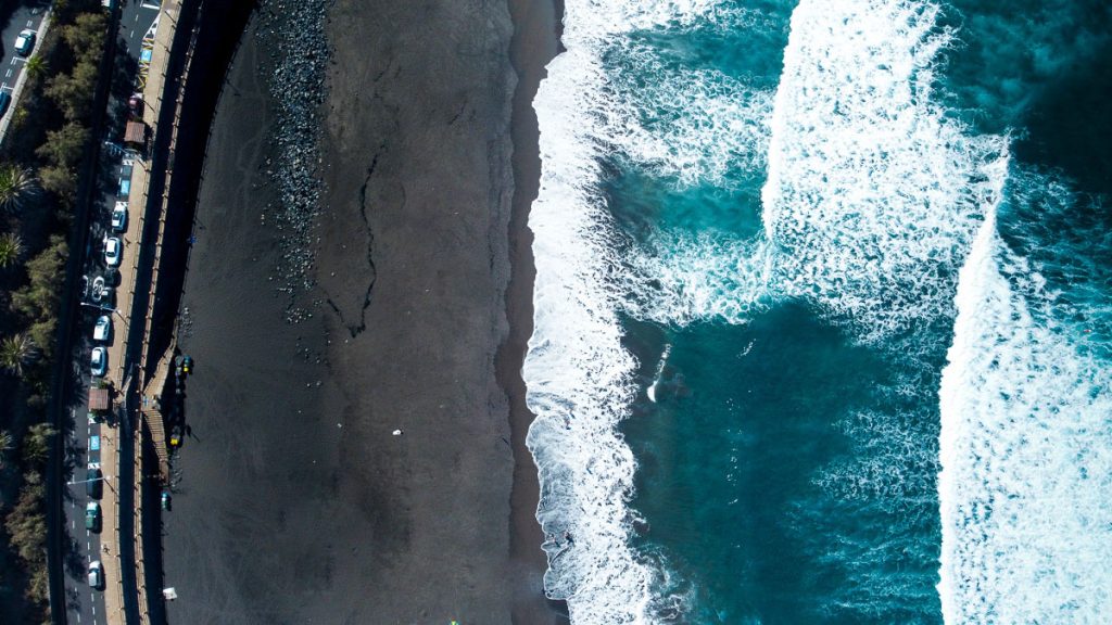 34 Incredible Weekend Trips From Barcelona 30 Aerial view of a unique black sand beach in Tenerife, Canary Islands, with the contrasting white surf of the waves meeting the shore next to a coastal road lined with cars and palm trees.
