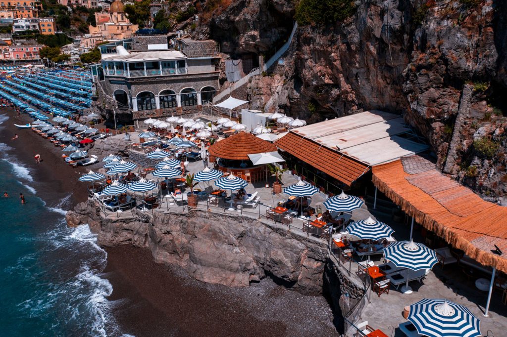 20 Best Amalfi Coast Beach Clubs 27 Aerial view of La Scogliera beach resort with striped blue and white umbrellas and a rustic terrace perched on the rocky Amalfi coast, overlooking a beach dotted with sunbathers.
