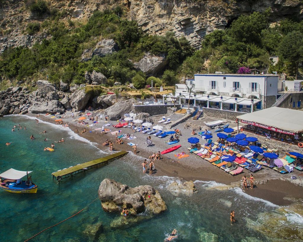 20 Best Amalfi Coast Beach Clubs 25 Vibrant scene at Laurito beach club featuring clusters of blue umbrellas on a pebbly shore, with a quaint white building nestled against a backdrop of steep, green-clad cliffs.