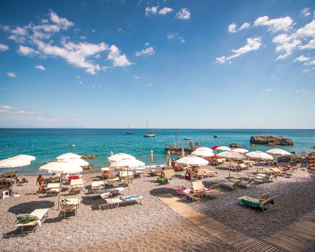 20 Best Amalfi Coast Beach Clubs 31 Lido degli Artisti in Amalfi under a cloud-speckled blue sky, featuring pristine white umbrellas and loungers on a pebbly beach, with the Mediterranean's azure waters stretching into the horizon.