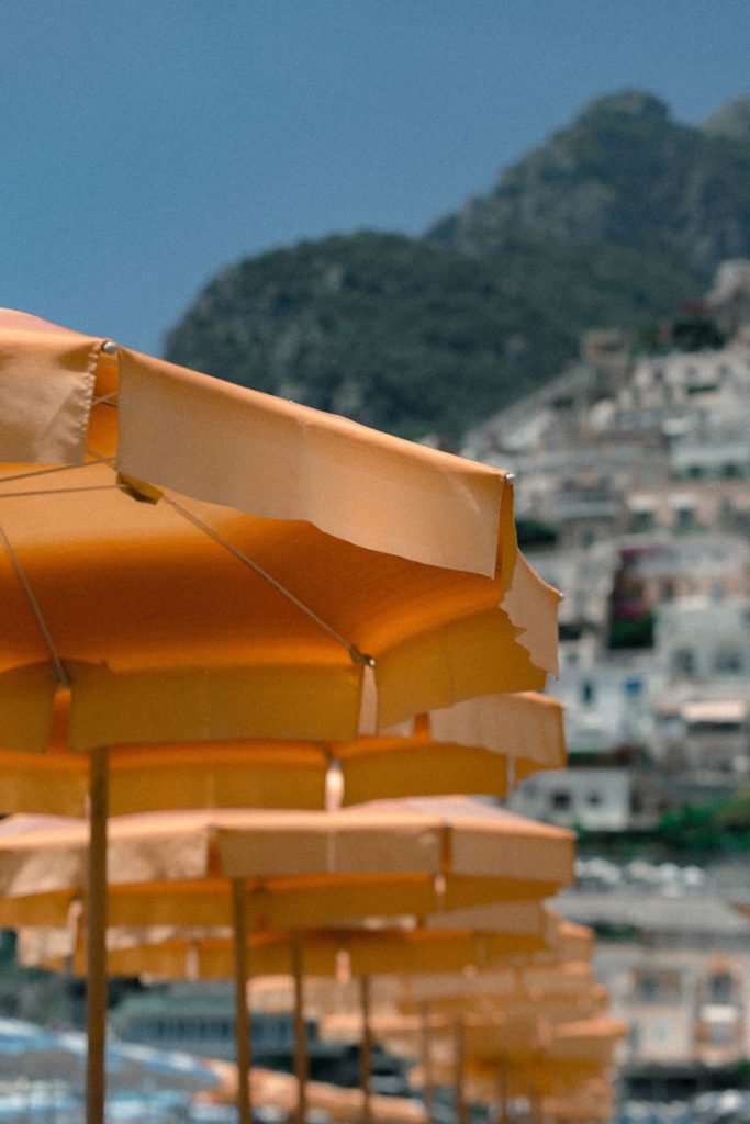 20 Best Amalfi Coast Beach Clubs 15 A lineup of vibrant orange beach umbrellas offering shade on a sunny day with a softly blurred background showcasing a mountainous landscape and the architecture of a coastal town.