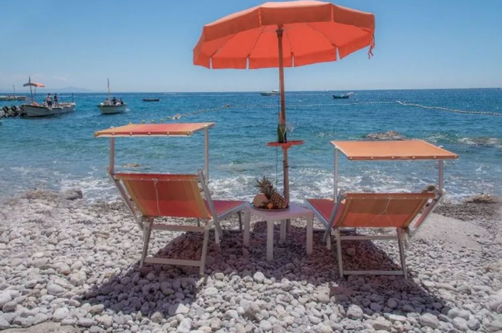 20 Best Amalfi Coast Beach Clubs 30 Seaside view of Santa Croce beach with red-orange umbrellas and matching sun loungers on a rocky shore, clear blue waters in the foreground, and boats leisurely passing by