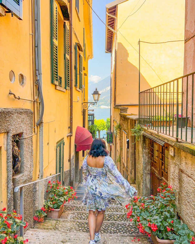 Bellagio Or Varenna: Where To Stay & Which Is Better? 29 woman walking down colorful orange and yellow homes in a narrow cobbled street in Bellagio, Lake Como