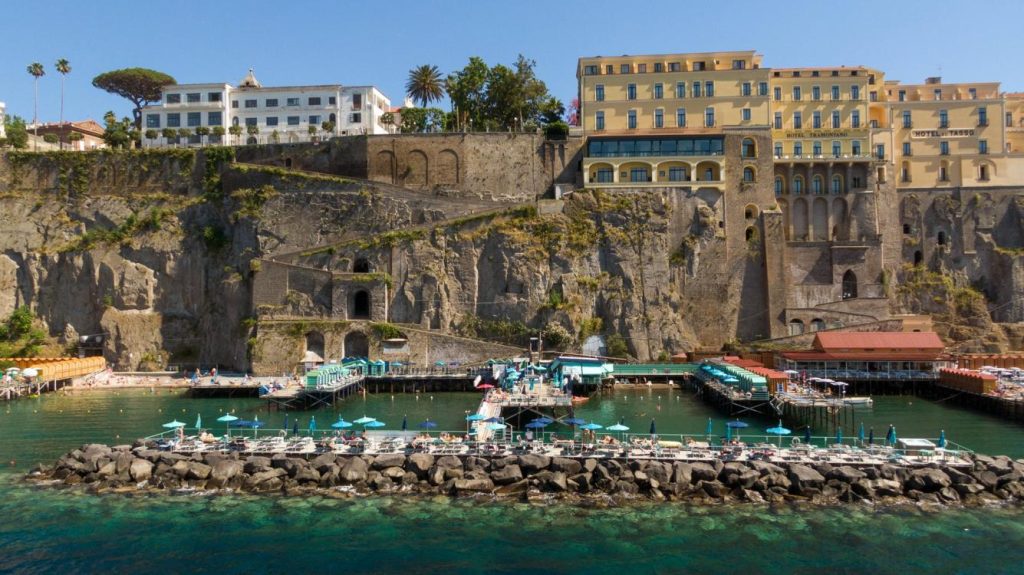 20 Best Amalfi Coast Beach Clubs 36 Marameo beach in Sorrento with white and teal umbrellas and sun loungers on a pier extending into the clear blue waters, backed by towering cliffs with lush greenery and historic buildings