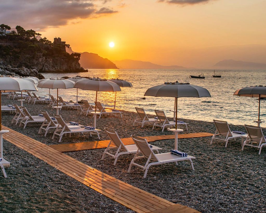20 Best Amalfi Coast Beach Clubs 32 Sunset at Marinella beach club where the sky meets the calm sea, with light grey umbrellas closed for the day and loungers inviting relaxation on the pebbly shore.