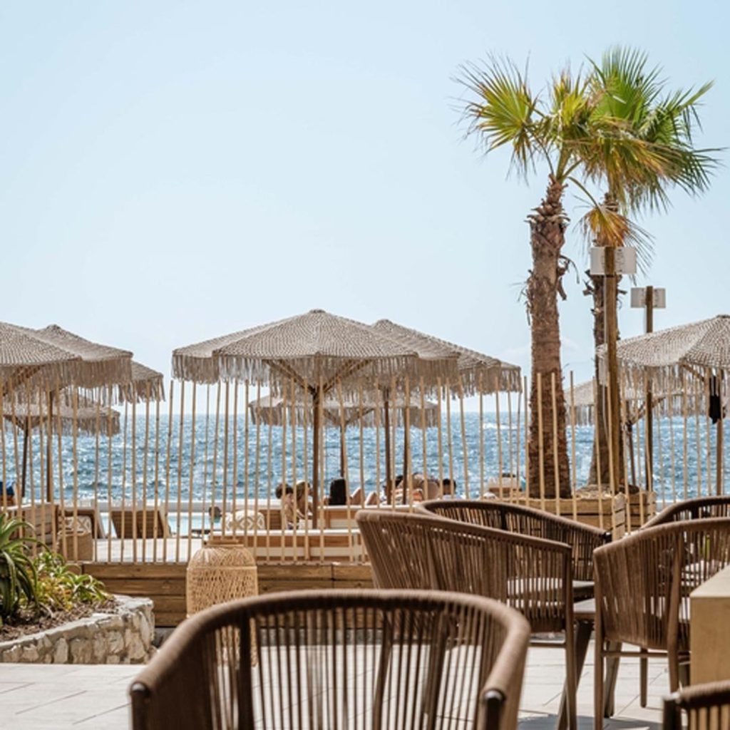 20 Best Amalfi Coast Beach Clubs 35 An elegant beachfront setting with chic wicker furniture under stylish straw umbrellas, a lone palm tree adding a tropical touch against the backdrop of a calm sea.