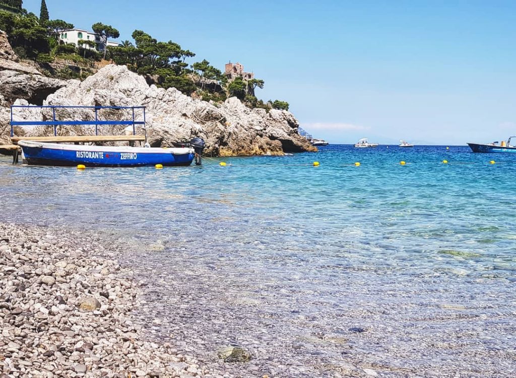 20 Best Amalfi Coast Beach Clubs 33 Clear turquoise waters lap against a pebble-covered shore with a blue boat named Ristorante Zeffiro moored nearby. In the background, rugged cliffs adorned with greenery and a quaint building overlook the serene Mediterranean setting.