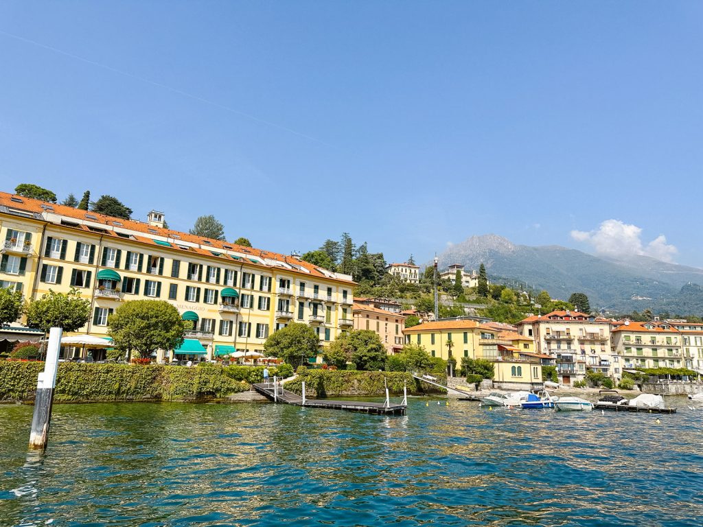 The Best Private Boat Tours Lake Como 14 Scenic view from Lake Como of the elegant Grand Hotel Menaggio, with its yellow faรงade and green shutters, set against a backdrop of lush hills under a clear blue sky.