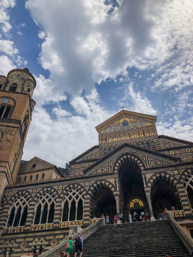 Positano Or Amalfi: Where To Stay & Which Is Better? 32 The ornate exterior of Amalfi Cathedral, featuring intricate striped arches, a grand staircase, and a bell tower under a partly cloudy sky.