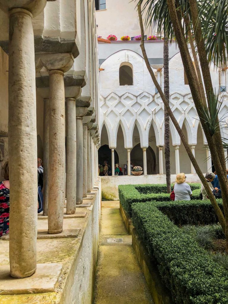 Positano Or Amalfi: Where To Stay & Which Is Better? 34 The Cloister of Paradise in Amalfi, showcasing a serene garden with trimmed hedges, stone columns, and an archway leading to an inner courtyard.