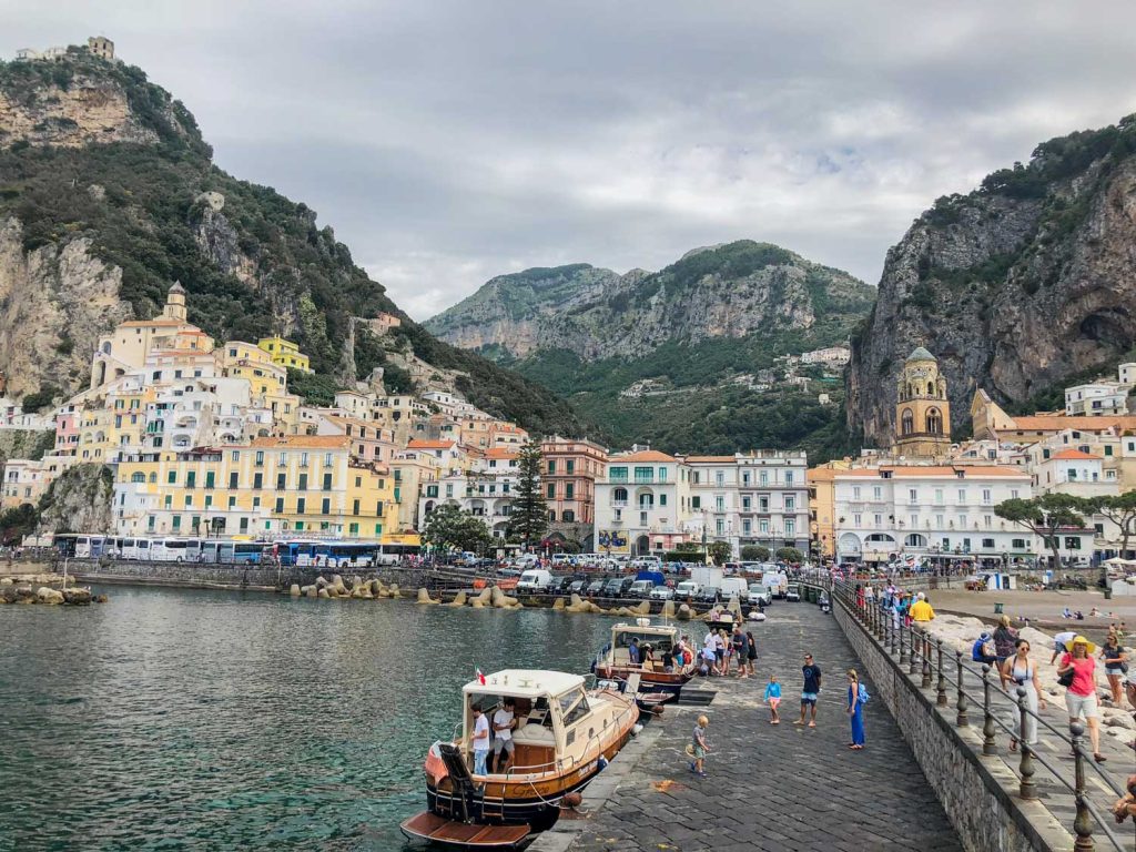 Positano Or Amalfi: Where To Stay & Which Is Better? 35 Amalfi as seen from Marina Grande, showcasing vibrant buildings cascading down the rugged hillside to the waterfront, with boats and people along the pier.