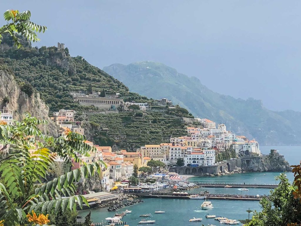 Positano Or Amalfi: Where To Stay & Which Is Better? 22 A panoramic view of Amalfi, featuring colorful buildings nestled on a lush hillside overlooking a serene blue sea with boats anchored in the bay.