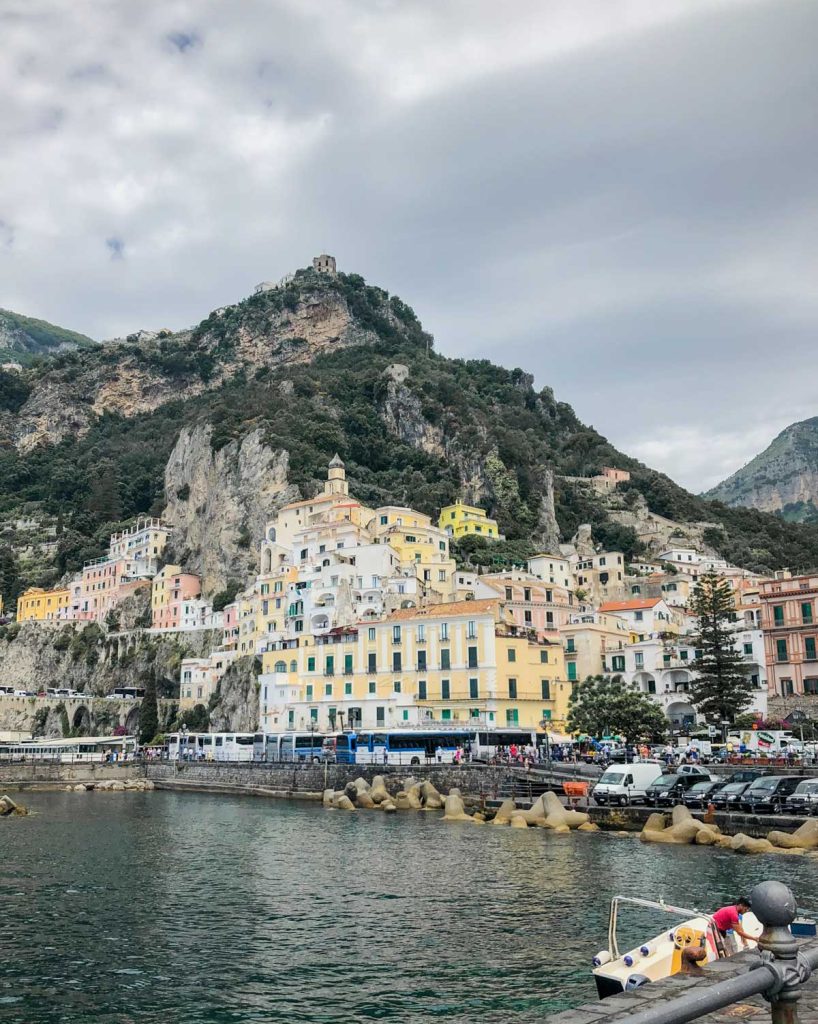 Positano Or Amalfi: Where To Stay & Which Is Better? 12 Scenic view of Amalfi from the Marina Grande, with charming multicolored buildings against a backdrop of steep cliffs and a calm turquoise sea in the foreground.