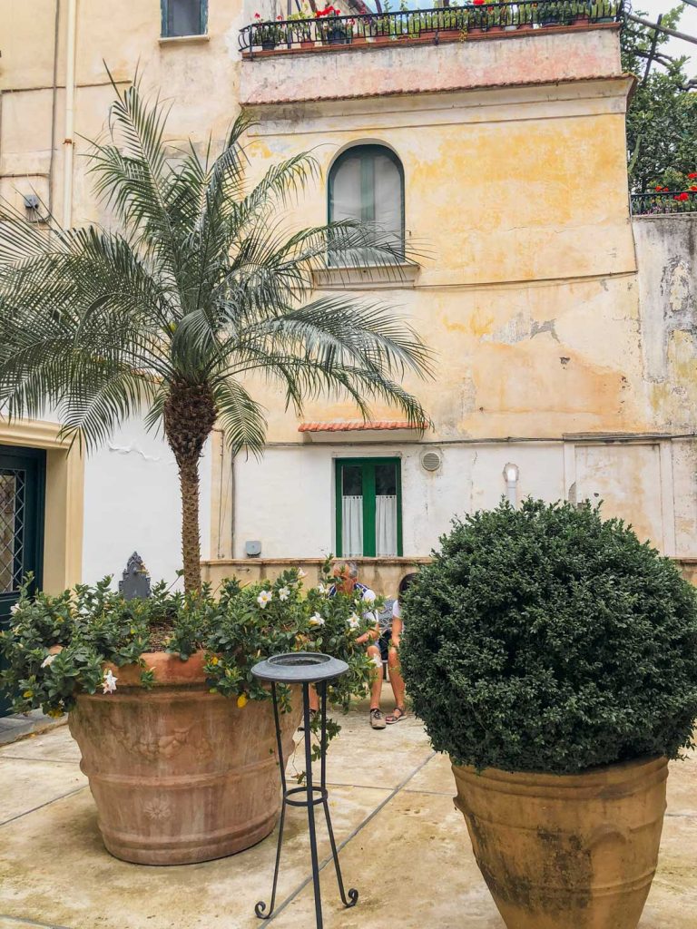 Positano Or Amalfi: Where To Stay & Which Is Better? 28 A relaxed scene showing Amalfi's lifestyle, with potted plants and a palm tree in front of an old building with pastel walls, where people sit and chat.
