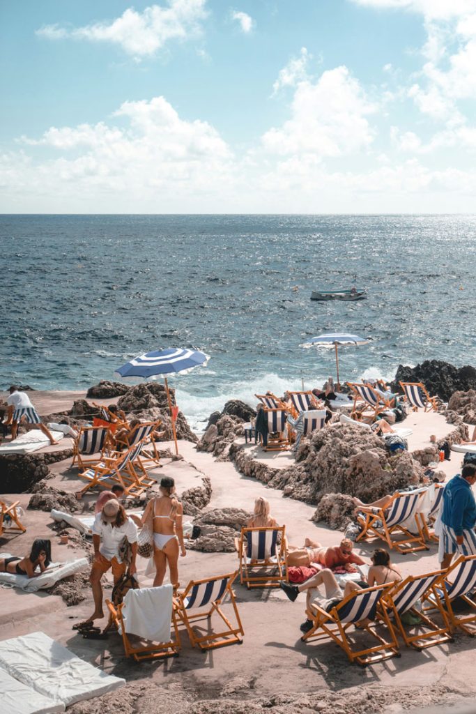 The Most Luxurious 5 Day Amalfi Coast Itinerary 21 A chic beach club on Capri's rocky shoreline featuring striped lounge chairs and umbrellas, with visitors relaxing near the crystal-clear Mediterranean waters.