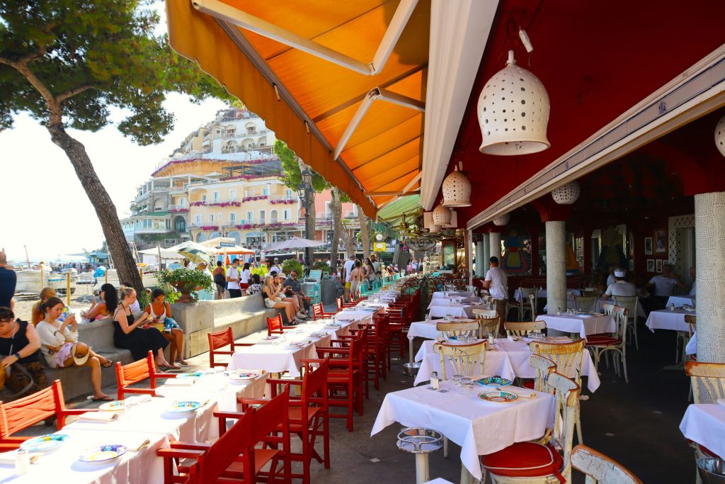 The Most Luxurious 5 Day Amalfi Coast Itinerary 41 A lively beachfront scene in Positano featuring vibrant restaurants with outdoor seating under orange awnings, overlooking the sea and the iconic hillside architecture.