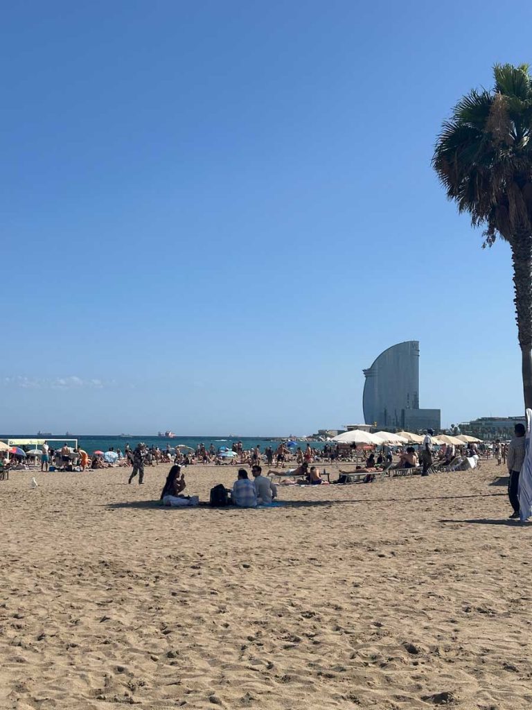 3 Days in Barcelona: The Perfect Itinerary for First-Time Visitors 38 A view of Barceloneta Beach, with the distinctive sail-shaped W Hotel in the background. The sandy shore is filled with beachgoers lounging under umbrellas, while palm trees frame the image.