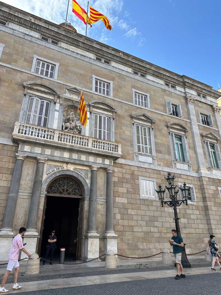 3 Days in Barcelona: The Perfect Itinerary for First-Time Visitors 53 A grand neoclassical building with Spanish and Catalan flags waving on the rooftop. The entrance is adorned with an arched doorway labeled "Palau de la Generalitat," with pedestrians and a security guard standing nearby.
