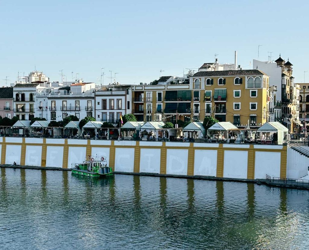 3 Most Amazing Days In Seville, Spain 32 Riverside view of the vibrant neighborhood of Triana in Seville, with rows of colorful houses, green-roofed stalls along the promenade, and a green boat floating in the river.