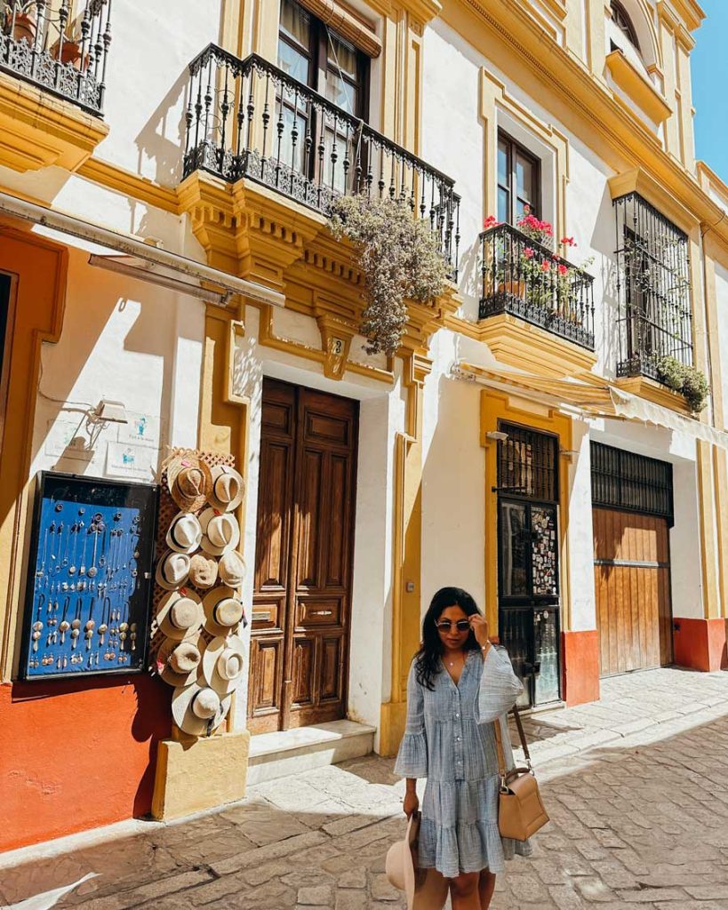 3 Most Amazing Days In Seville, Spain 37 Woman in a light blue dress walks past a souvenir stall in Seville’s Barrio Santa Cruz, with sun hats and trinkets on display against a vibrant white and mustard-colored building.