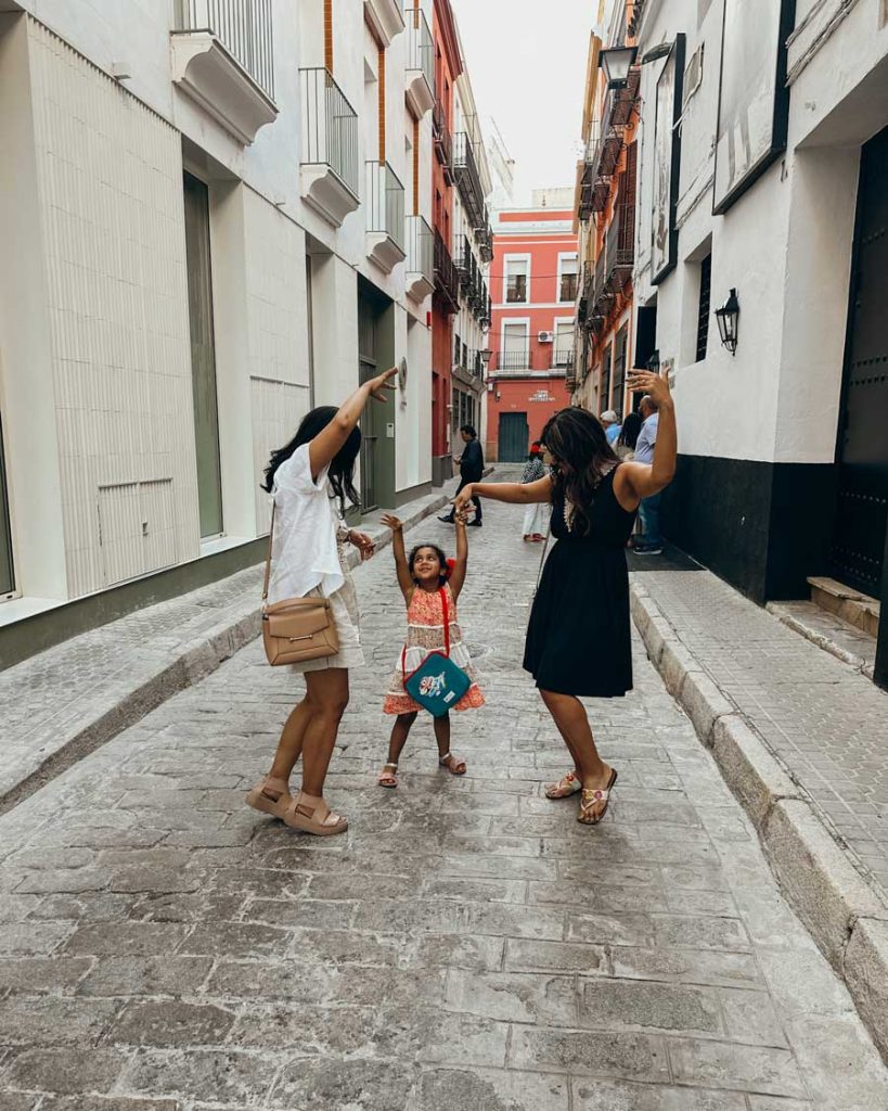3 Most Amazing Days In Seville, Spain 27 Two women and a young girl joyfully dance together in a narrow residential street in Seville, surrounded by colorful facades and hanging laundry in the background.