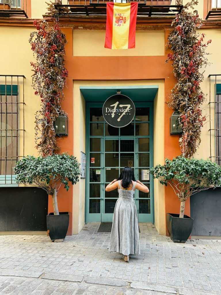 3 Most Amazing Days In Seville, Spain 33 A woman in a checked dress walks toward the entrance of Hotel Doña Maria in Seville, with a Spanish flag above the teal door and dried floral decor framing the doorway.