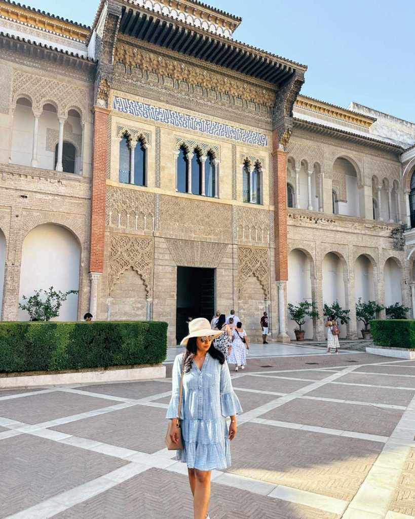 3 Most Amazing Days In Seville, Spain 11 Woman in a striped blue dress and sunhat walks through the main courtyard of the Royal Alcázar in Seville, standing in front of a stunning Mudejar-style facade with intricate carvings and arched windows.