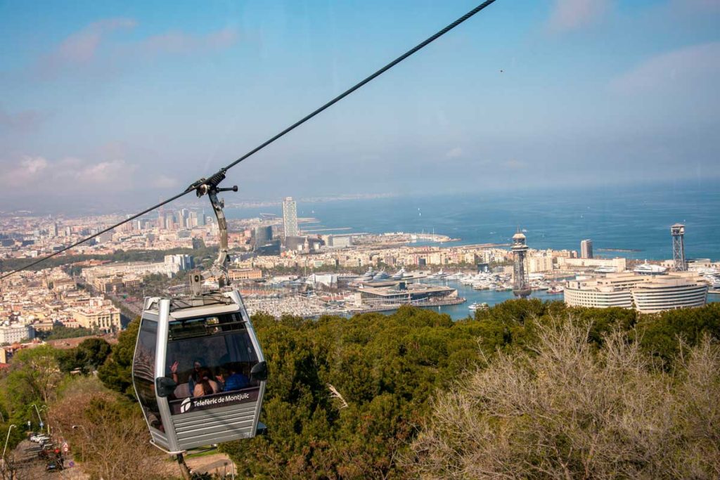 3 Days in Barcelona: The Perfect Itinerary for First-Time Visitors 37 A breathtaking view of Barcelona from the Montjuïc Cable Car, with a gondola labeled 'Telefèric de Montjuïc' suspended over lush green hills. The cityscape stretches toward the Mediterranean Sea, featuring the bustling Port of Barcelona, modern high-rises, and iconic towers. The blue sky and distant horizon complete the panoramic scene.