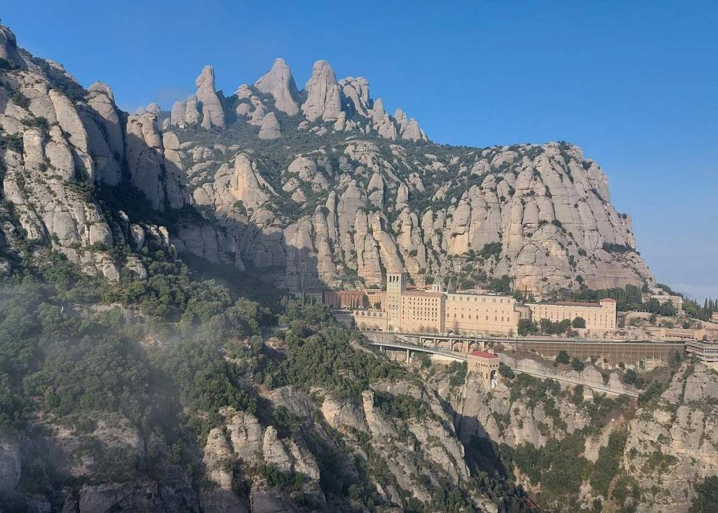 34 Incredible Weekend Trips From Barcelona 13 A stunning view of Montserrat, Spain, featuring the famous Santa Maria de Montserrat Abbey nestled among dramatic, uniquely shaped rock formations. The jagged mountain peaks rise in the background, partially covered with green vegetation, while the monastery's beige stone buildings stand out against the rugged terrain. A railway and roads are visible, connecting the site to the surrounding area under a clear blue sky.