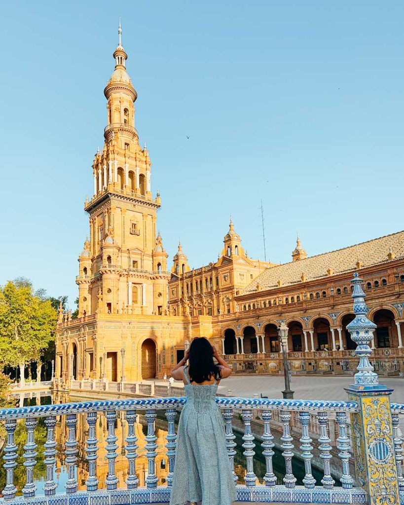3 Most Amazing Days In Seville, Spain 10 A woman in a light blue gingham dress stands by a tiled balustrade, looking at the golden-hued towers and arched colonnades of Plaza de España glowing in the morning sun.
