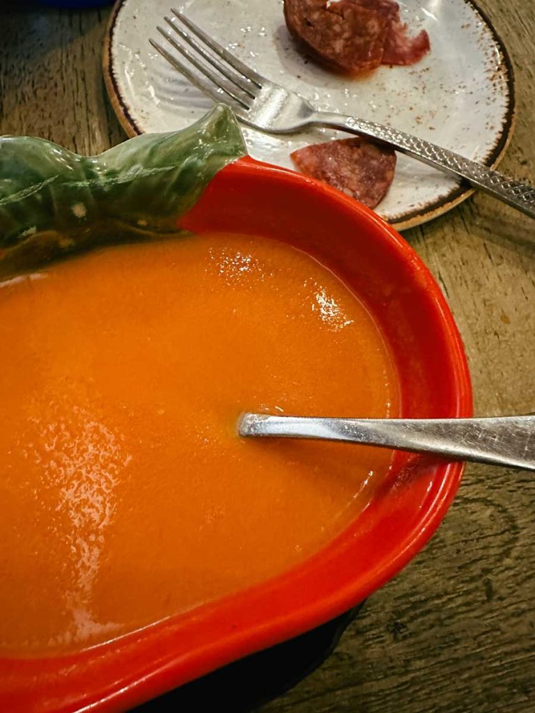 3 Most Amazing Days In Seville, Spain 34 A vibrant orange bowl of gazpacho served in a decorative leaf-shaped dish, next to a plate with slices of sausage and a fork.