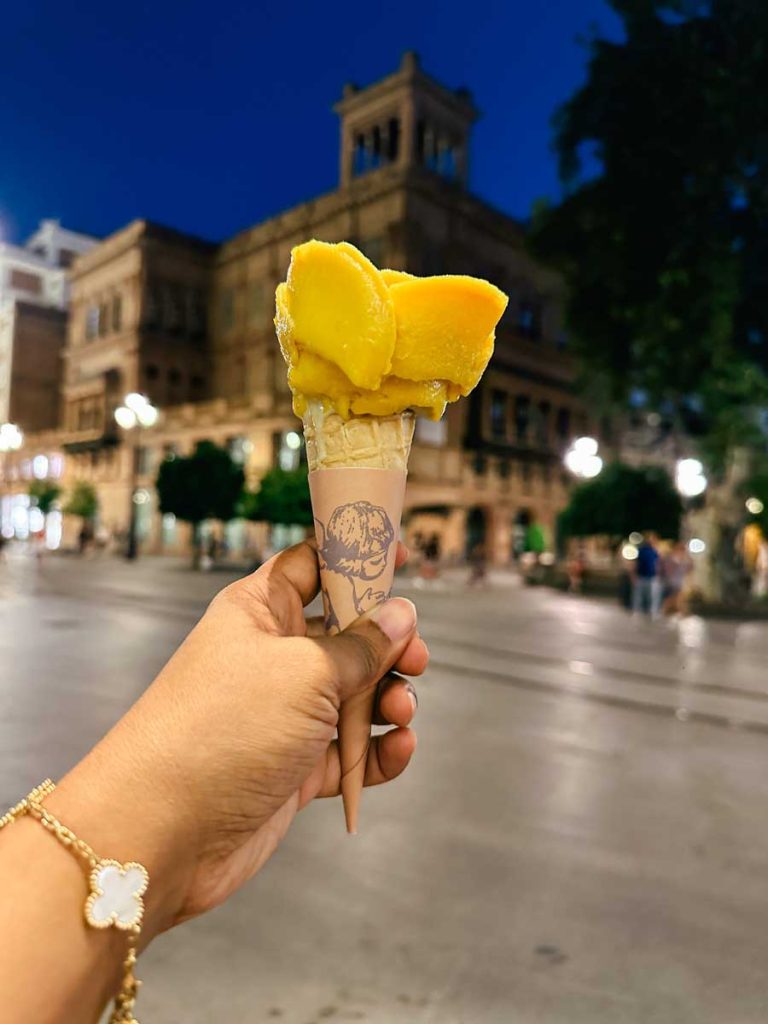 3 Most Amazing Days In Seville, Spain 15 Close-up of a hand holding a rose-shaped mango gelato cone from Amarino, with a softly lit plaza and historic building in the background.