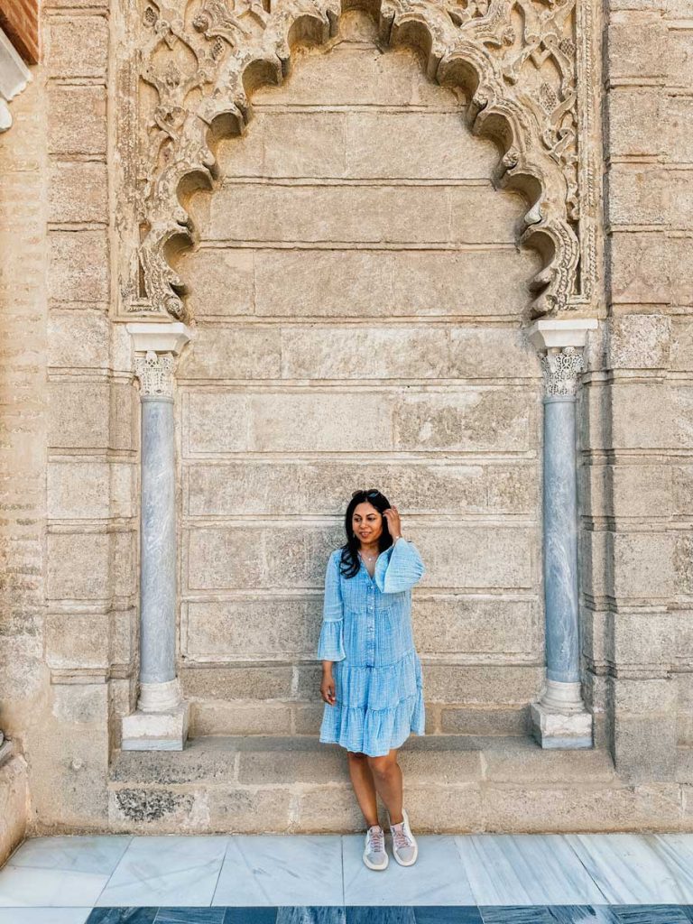 3 Most Amazing Days In Seville, Spain 38 Woman in a blue dress poses beneath a stone horseshoe arch with intricate carvings at the Royal Alcázar of Seville, framed by columns and geometric tile flooring.