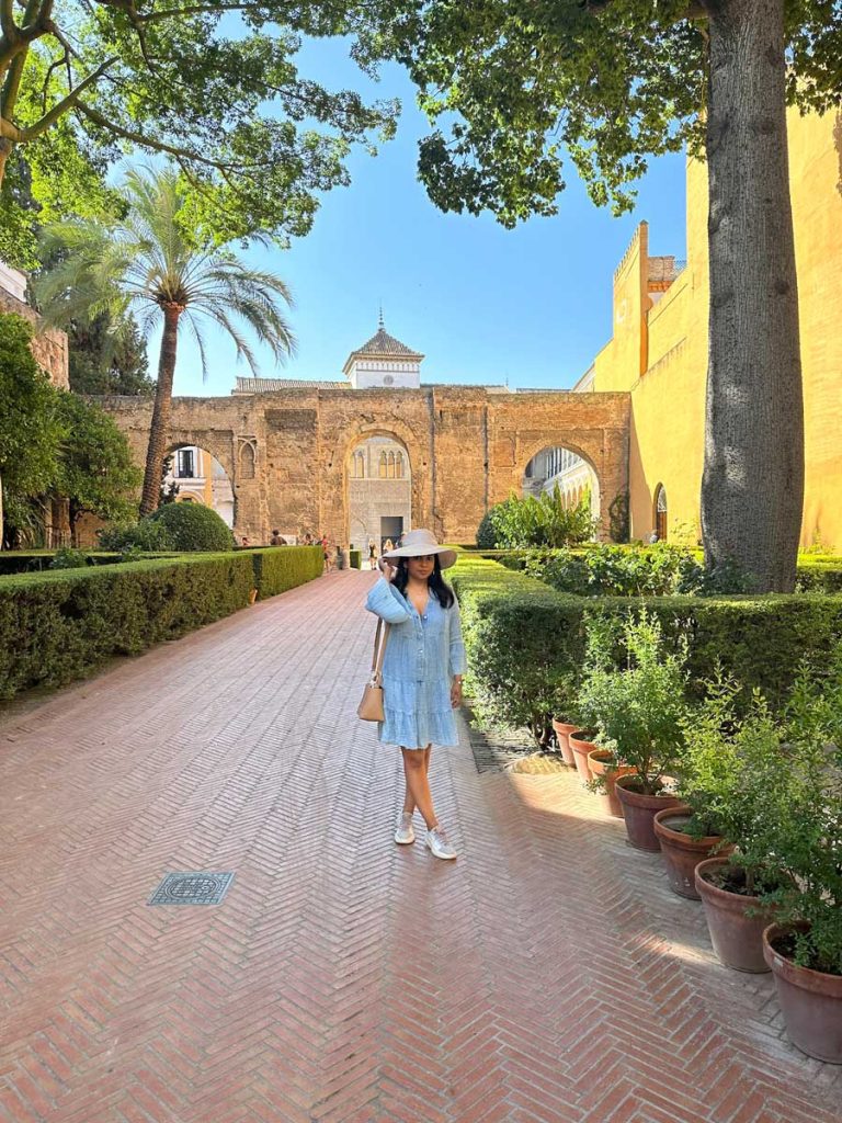 3 Most Amazing Days In Seville, Spain 16 A woman in a light blue dress and sunhat walks down a garden path lined with terracotta pots, toward the grand gate of the Royal Alcázar in Seville.