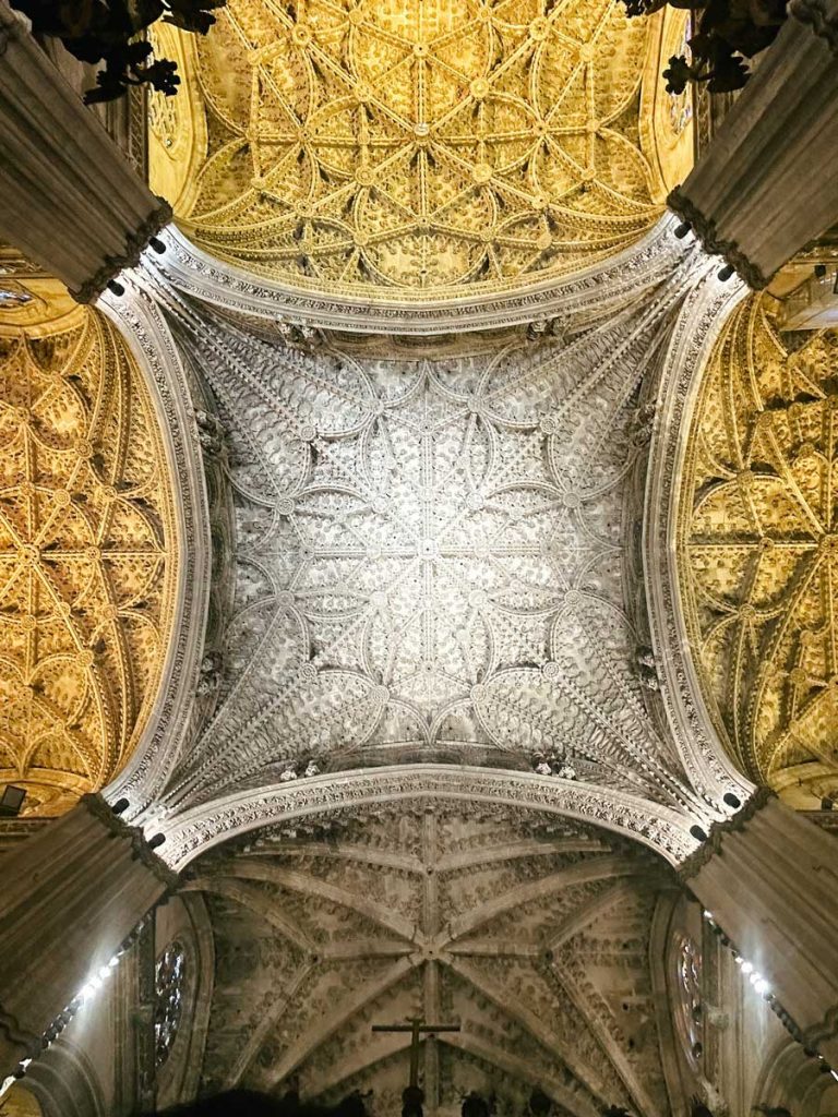 3 Most Amazing Days In Seville, Spain 21 Intricate ribbed Gothic vault ceiling inside Seville Cathedral, showcasing golden and cream-toned details in a symmetrical floral pattern.