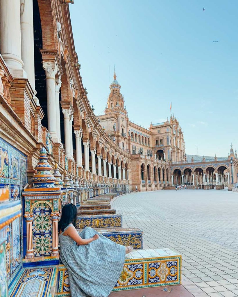20 Best European Summer Destinations To Visit This Year 31 Woman in a gingham dress sitting on the colorful tiled benches at Plaza de España in Seville, surrounded by curved colonnades and historic towers.