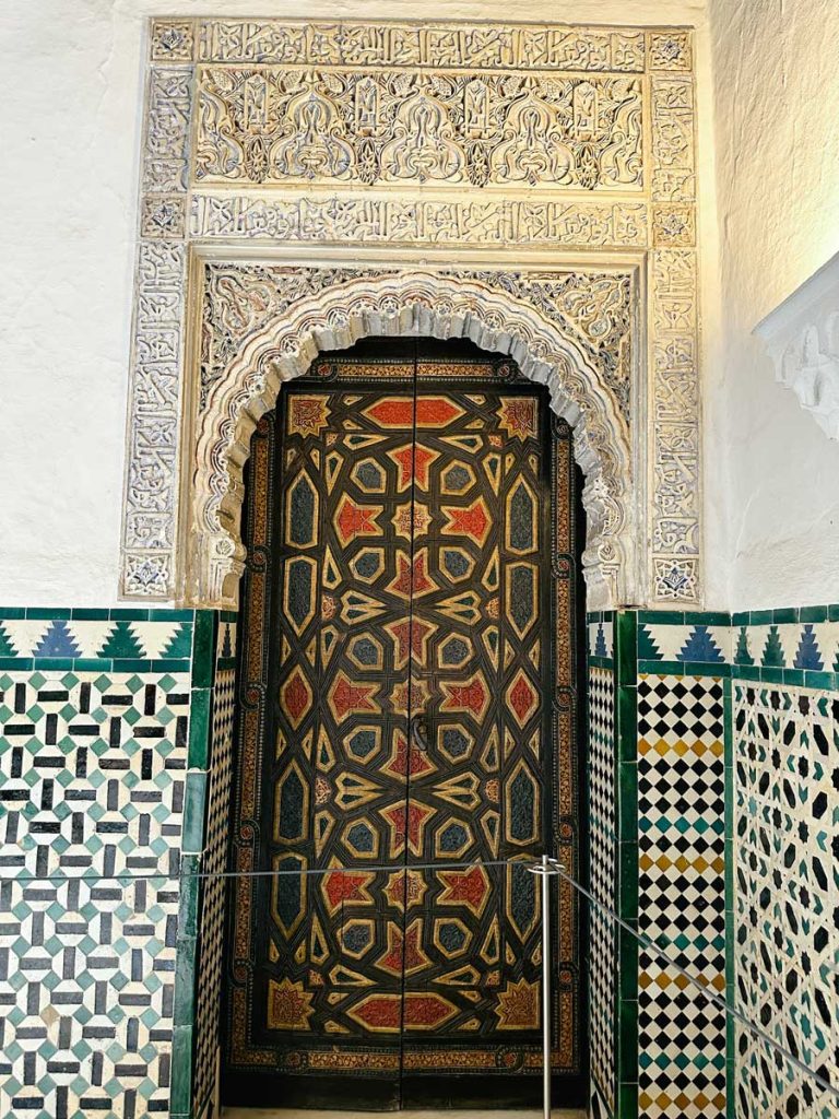 3 Most Amazing Days In Seville, Spain 18 Close-up of an elaborately carved Moorish-style doorway at the Royal Alcázar of Seville, framed by detailed tile work and Arabic calligraphy with a colorful geometric wooden door.