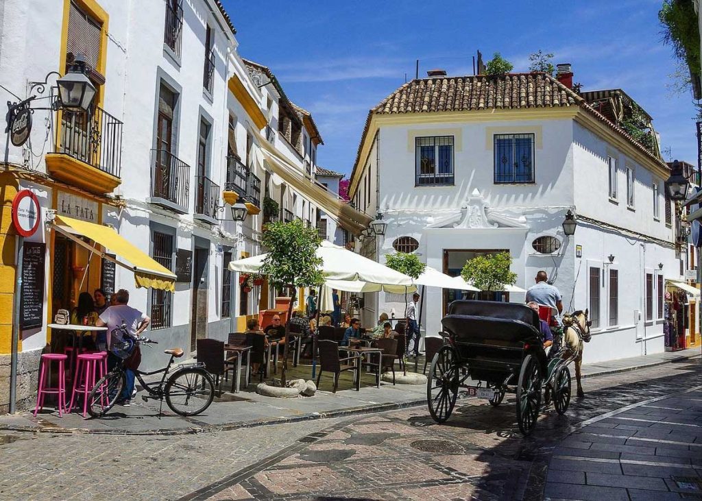3 Most Amazing Days In Seville, Spain 31 Charming street in Córdoba, Spain, lined with whitewashed buildings and yellow trim, featuring outdoor café seating, a horse-drawn carriage, and a bicycle parked outside "La Tata" bar under sunny blue skies.