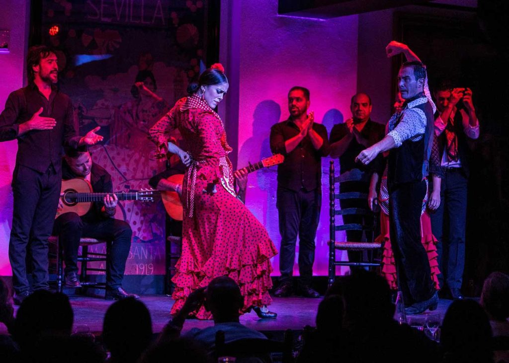 3 Most Amazing Days In Seville, Spain 28 Vibrant flamenco performance in Seville featuring a woman in a red polka dot dress dancing passionately, surrounded by clapping musicians and performers against a purple-lit backdrop.
