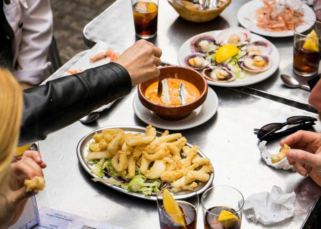 3 Most Amazing Days In Seville, Spain 36 Overhead shot of a lively tapas table in Seville with plates of seafood, fried calamari over salad, gazpacho, and glasses of cola garnished with orange slices.