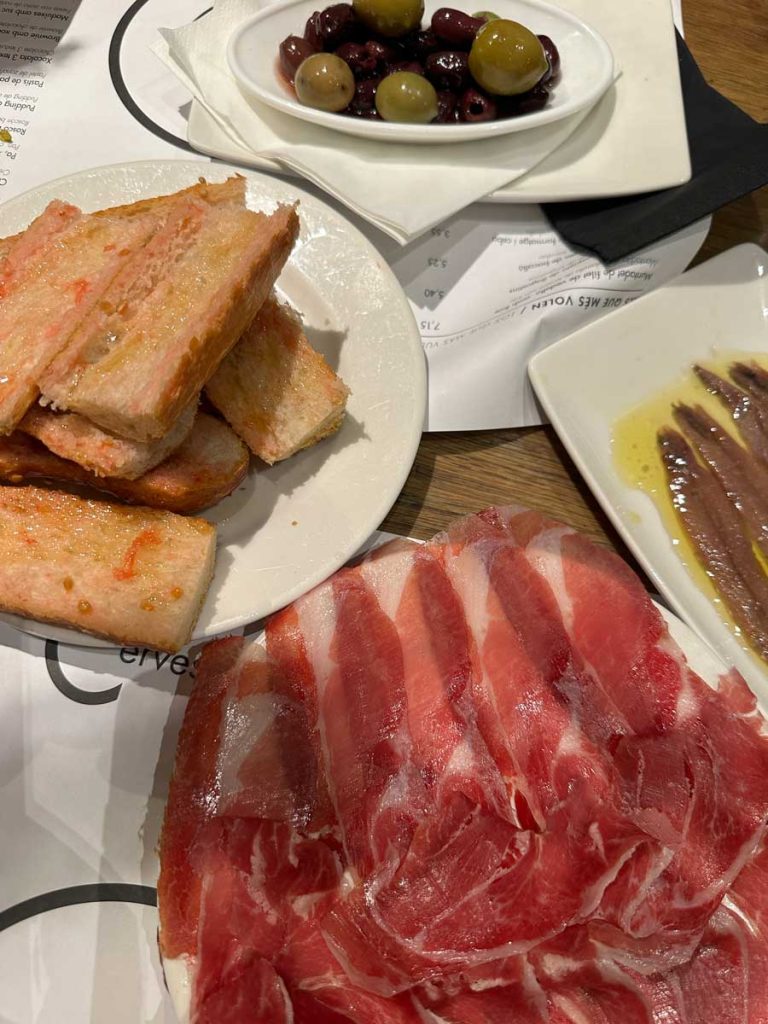 3 Days in Barcelona: The Perfect Itinerary for First-Time Visitors 43 A close-up of a tapas spread, including thinly sliced jamón ibérico, crispy pan con tomate, a plate of marinated olives, and anchovies drizzled with olive oil. The dishes are served on a table with a menu and napkins underneath.