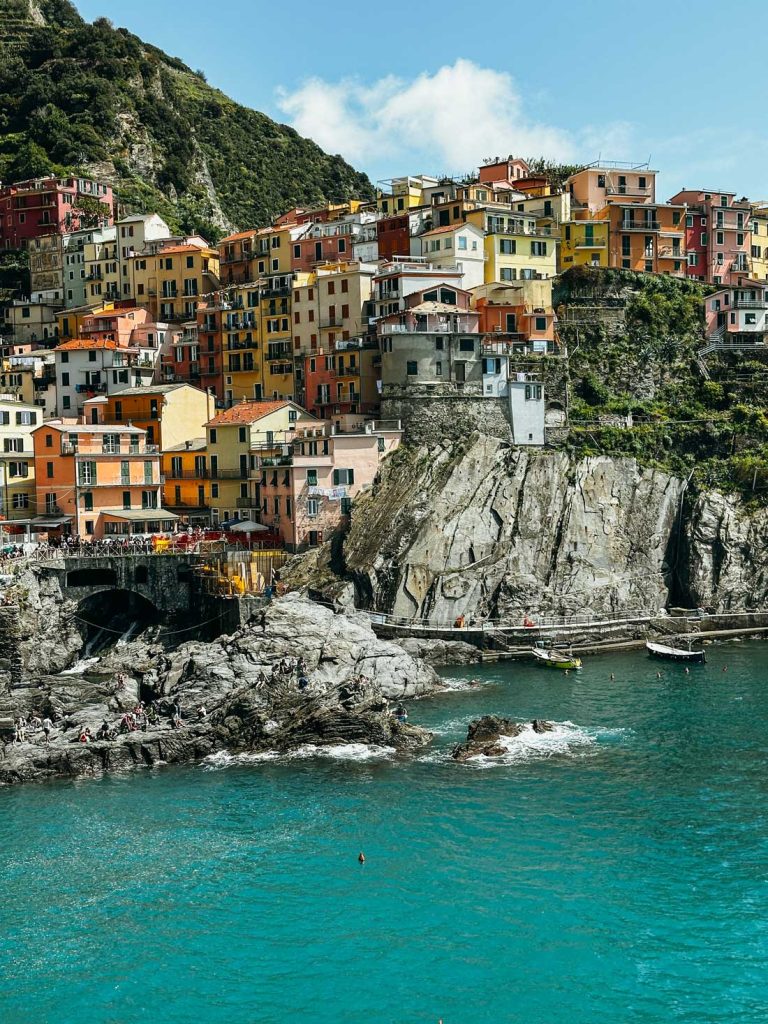 20 Best European Summer Destinations To Visit This Year 13 The colorful cliffside village of Manarola in Cinque Terre overlooks turquoise waters and rugged rocks dotted with visitors.